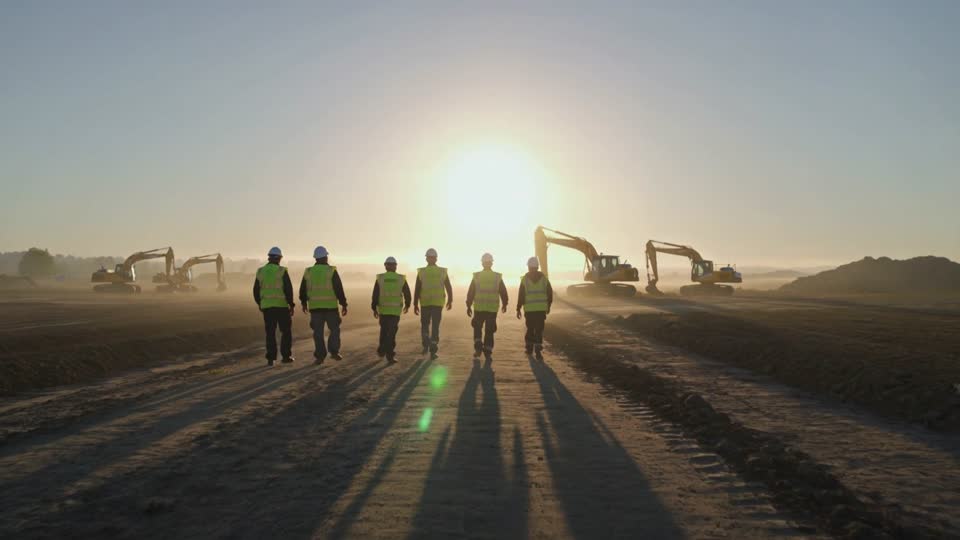A crew in hi-vis vests walking across a graded site toward their machines at sunrise